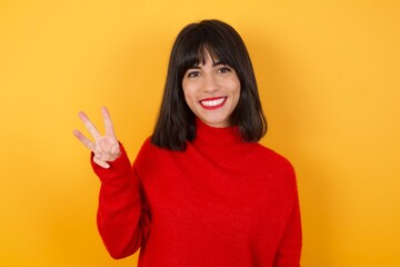 Caucasian brunette woman wearing red casual sweater isolated over yellow background showing and pointing up with fingers number three while smiling confident and happy.