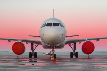 Front view of white airplane. Jet commercial aircraft on airport apron, morning sunrise orange red sky, rain puddles. Modern technology in fast transportation, private business travel, charter flights