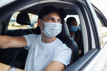 Fototapeta na wymiar Young taxi driver looks out of a car's window while driving through the city with a passanger wearing a medical mask. Business trips during pandemic, new normal and coronavirus travel safety concept.