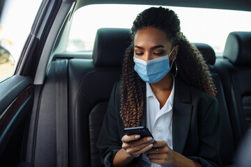 Young business woman in a mask checking her mobile cell phone on a backseat of a taxi during covid-19 quarantine. Business trips during pandemic, new normal and coronavirus travel safety concept.