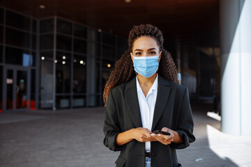 Young businesswoman wearing a medical mask stands near the office center. Officially looking girl with a phone in her hands waiting outside. Leading business during Covid-19 pandemic concept.