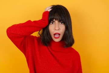 Embarrassed Caucasian brunette woman wearing red casual sweater isolated over yellow background with shocked expression, expresses great amazement, Puzzled woman poses indoor