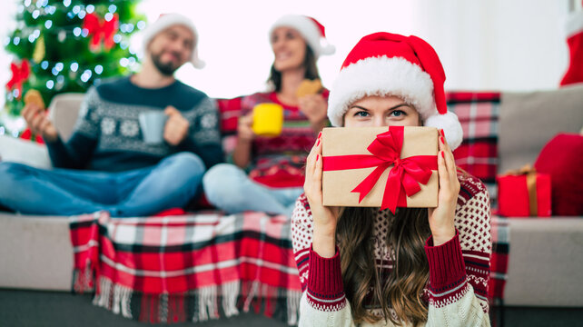 Close up photo of beautiful young happy smiling woman in a Christmas sweater and Santa hat is holding a gift box in hands at home on the background of her friends and Christmas tree.