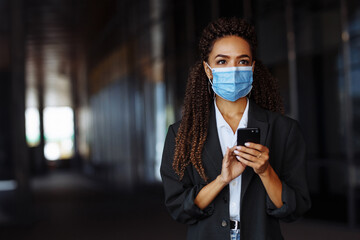 Young businesswoman wearing a medical mask stands near the office center. Officially looking girl with a phone in her hands waiting outside. Leading business during Covid-19 pandemic concept.