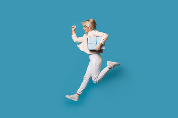 Caucasian woman running with a present on a blue studio wall wearing white clothes and blonde hair