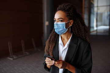 Young businesswoman wearing a medical mask stands near the office center. Officially looking girl with a phone in her hands waiting outside. Leading business during Covid-19 pandemic concept.