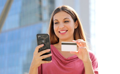 Close up of delighted nice woman holding credit card and using smartphone outdoors