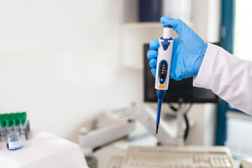 Laboratory technician holding pipette tool in his hand in blue protective glove. Automatic micropipette with computer monitor, keybord and automated lab analyser in white background. Selective focus.