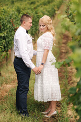 Fototapeta premium Groom and bride walking in vineyard in their wedding day. Blond hair woman in white dress hold wedding bouquet in hands