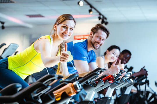 Woman showing thumbs up sign exercising of fitness bike in gym - Powered by Adobe