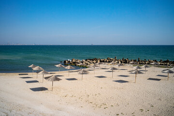 Late September empty beach in a Romanian resort at the Black Sea with straw umbrellas and rocky edges.