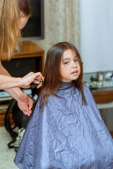 Portrait of a young girl getting her hair cut in home. Womans hand with hairdressing scissors. Little child sits patiently while having her haircut.