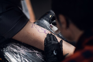 Close-up photo of a girl tattoo artist putting a tattoo on the arm, of a young guy in a tattoo...