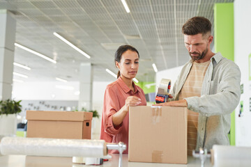 Waist up portrait of modern adult couple packing cardboard boxes with tape gun in storage facility or shipping service, copy space