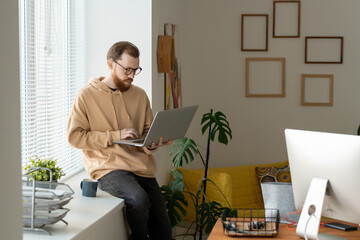 Young bearded freelancer using laptop while networking on windowsill