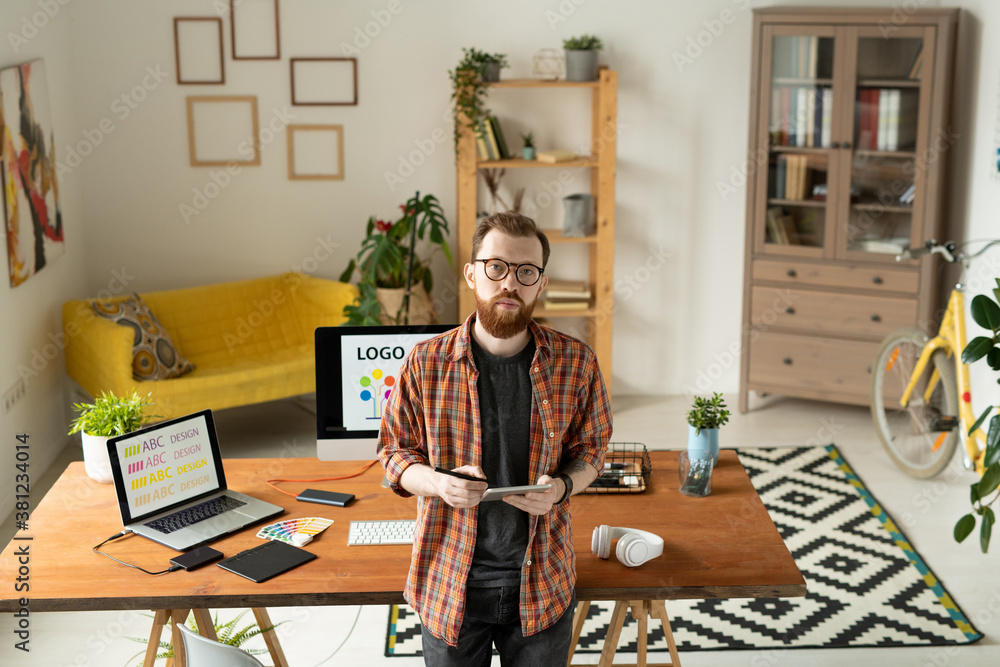 Wall mural serious creative designer standing by table with computer monitor on background