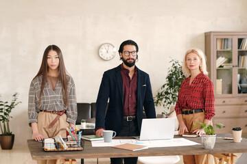 Three intercultural colleagues in smart casualwear standing in row along table