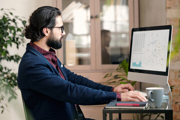 Serious young broker or economist in formalwear sitting in front monitor