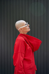 Cool fashion bald young woman wearing trendy red hoodie on backwards and sunglasses standing on...
