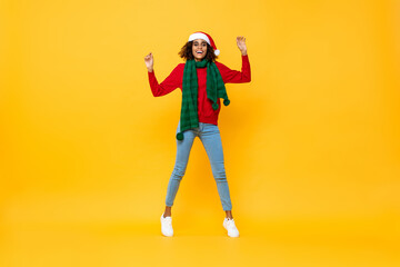 Fototapeta na wymiar Excited pretty woman in Christmas attire raising hands up in isolated yellow studio background
