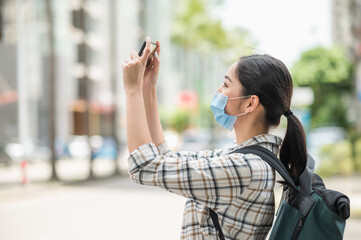 Young Asian beautiful woman travelers use her mobile phones to take the photo of view urban city downtown. She wearing protection mask for new normal safety travel trips.