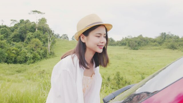Happy young Asian woman wearing a hat and smiling with keep luggage behind the car, travel in the summer on a clear day on green meadow background.