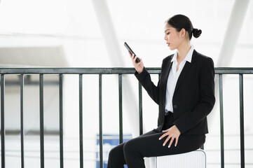 Young asian business woman sitting on trolley and using smartphone. Beautiful female in black suited.