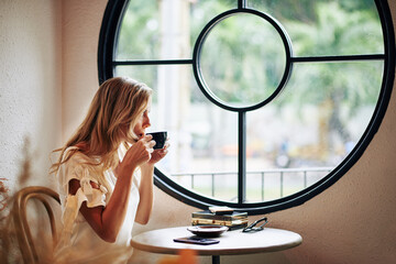 Beautiful young blond woman drinking cup of cappuccino and looking through cafe window