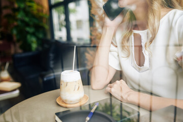 Cropped image of young woman drinking delicious sweet iced coffee in restaurant and calling on phone