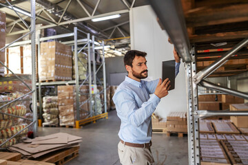 Young attractive bearded supervisor standing next to shelves in warehouse and checking on goods while holding and looking at tablet.