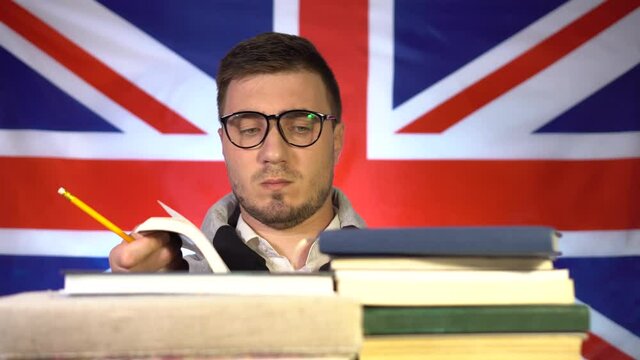 Cute guy student with glasses fluently leafs through a book from a pile of books on the background of the flag of Great Britain. Education in the UK. Education concept.