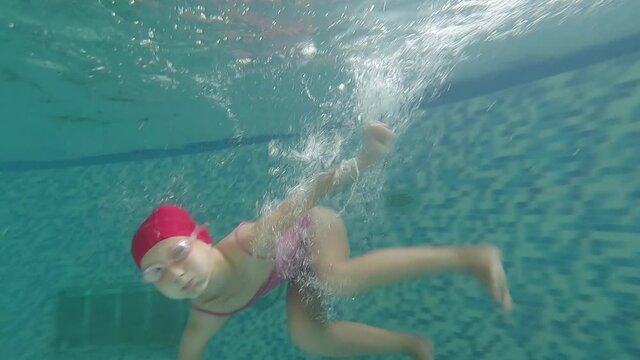 Funny little girl fooling around in the pool underwater and smiling. A child in a red bathing suit, cap and goggles dives and swims