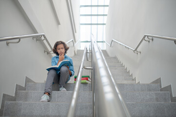 Schoolboy sitting on stairs, reading book, holding his chin
