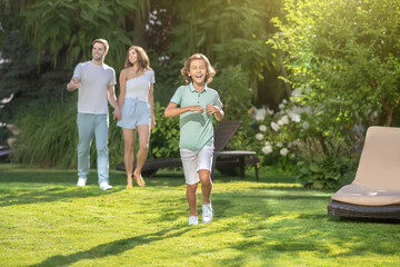 Obraz premium Smiling boy running on lawn, his parents walking behind