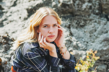 Thinking young blonde woman in blue dress sitting on stones on background of mountains