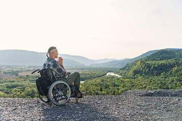Likable incapacitated young guy sitting alone in wheelchair on the hill on beautiful nature background and praying.