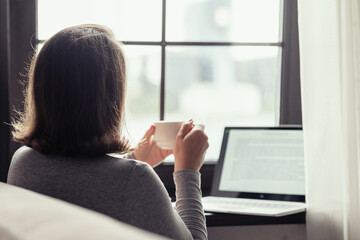 Back view of lonely woman enjoying having breakfast with cup of coffee working on laptop sitting...