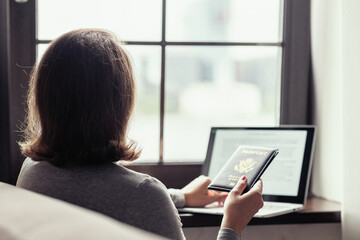 Woman holding american passport using laptop for making order and booking sitting near window. Online service. concept.