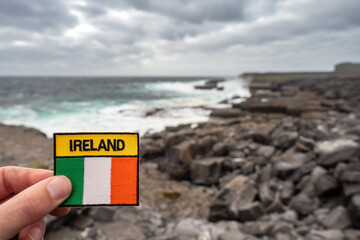 Tourist holding badge with sign Ireland and Irish National flag in focus. Rough stone coast line...