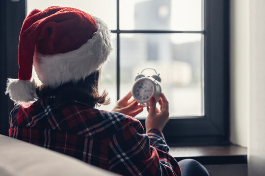 Back view of lonely sad young woman in a red santa claus christmas hat sitting near window and holding alarm clock. Concept