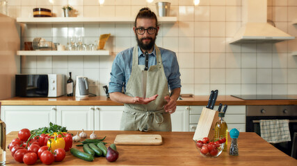 Young man, Italian cook in apron looking at camera, holding knife the tip of the blade while preparing healthy meal with vegetables in the kitchen. Cooking at home concept