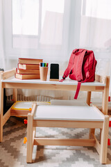 Fototapeta na wymiar School supplies on the school desk. Red backpack, white headphones, notebook, big red books, pens in the jar lay on the white school desk.