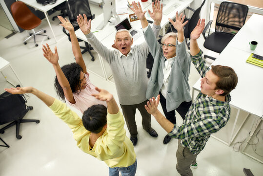 Group of diverse employees raising hands in the air, hoping for success on their plans while standing in the office, Aged woman and man, senior interns having first day at work