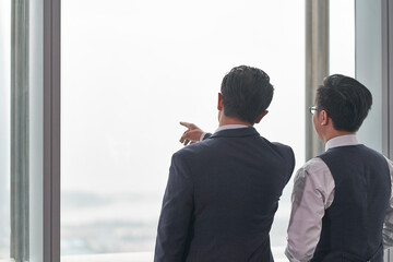 rear view of two asian businessmen standing in front of window talking discussing chatting...