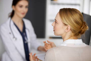 Fototapeta premium Friendly smiling female doctor and patient woman discussing current health examination while sitting in clinic. Medicine and healthcare concept