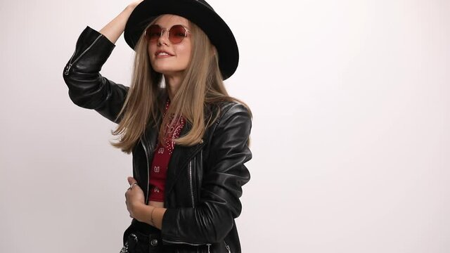 boho chic young woman wearing leather jacket and sunglasses, fixing hat and hair, smiling and laughing, moving and having fun, holding hands on hips and posing on grey background