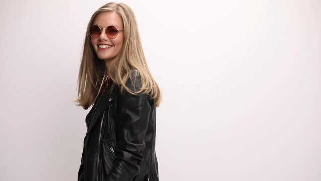 cool young casual girl wearing leather jacket and sunglasses arranging hair, smiling, moving in a side view position and posing, adjusting jacket on grey background