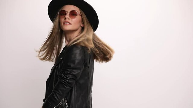 hippie young girl wearing leather jacket and sunglasses, arranging hat, moving in a back view position, looking down and smiling on grey background
