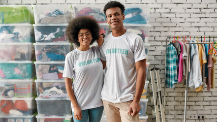 Two young diverse volunteers smiling at camera, posing together in front of rack and boxes full of...