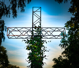 Christian steel cross structure with wild plants growing on it, blue evening skies and trees in the...